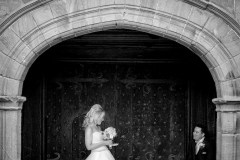The groom is seated in the doorway of Speke Hall. The bride is looking on. This photo is black & white.